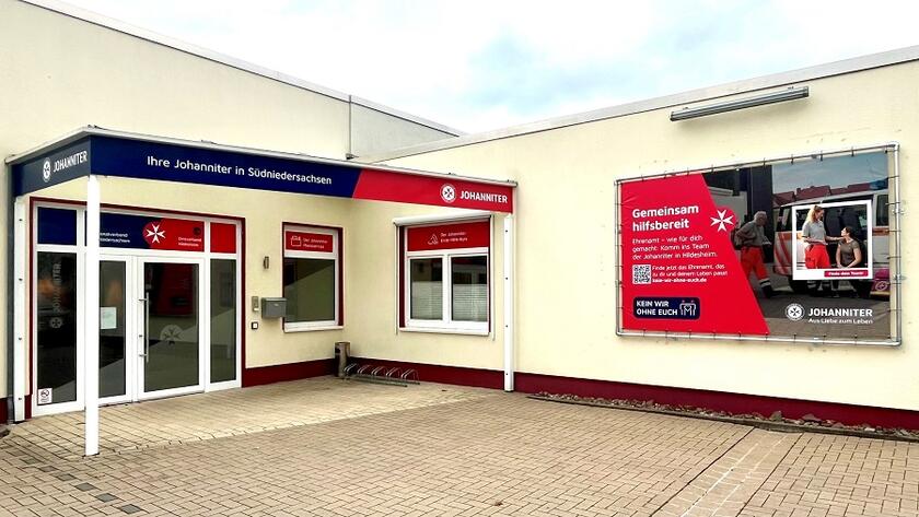 Ein gelbes Flachdachgebäude mit einer doppelseitiges gläsernen Eingangstür die überdacht ist an der rechten Hauswand ein Plakat mit Johannitern drauf.