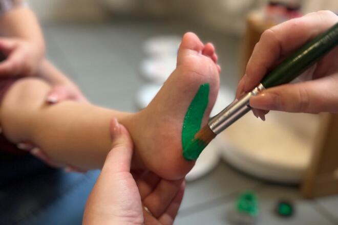 Eine Erzieherin der Kinderkrippe bemalt die Fusssohle eines Kindes mit grüner Farbe für einen Fußabdruck