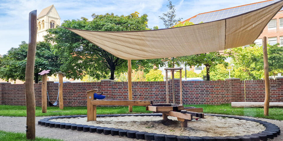 Ein Sandkasten mit Wasserspiel und einer Überdachung aus beigem Stoff. Im Hintergrund eine rote Backsteinmauer und Gebäude.