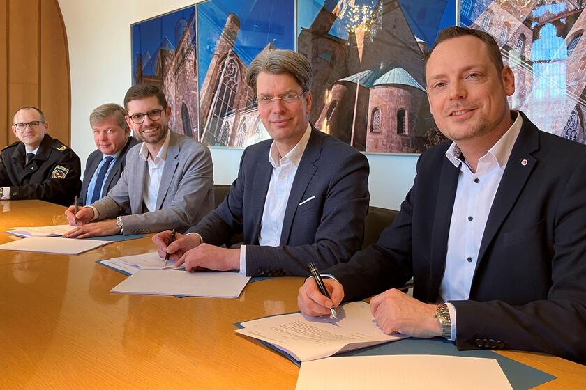 Fünf Männer an einem Konferenztisch. Drei haben Verträge vor sich und einen Stift in der Hand