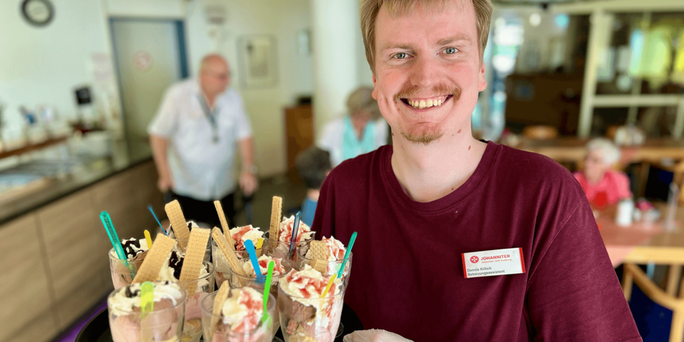 Ein junger Mann in einem dunkelroten T-Shirt mit Johanniter-Namensschild trägt ein Tablett mit Eisbechern
