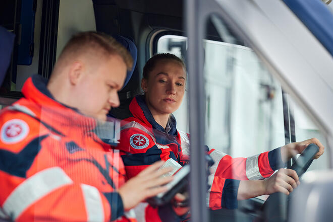 Zwei Johanniter Rettungskräfte im Einsatzfahrzeug, einer am Tablet, die andere fährt.