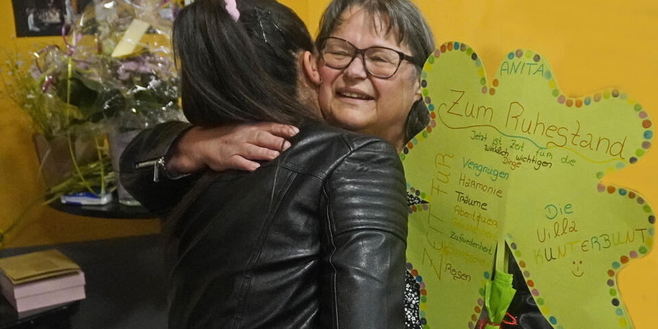 Eine Frau wird von einer anderen Frau umarmt und hält ein Papierkleeblatt mit Innschrift