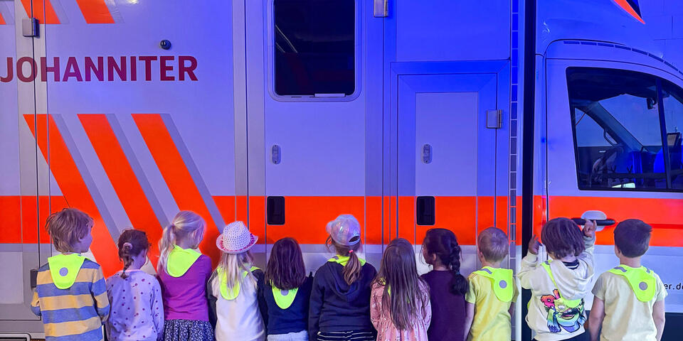 Kinder vor einem Rettungswagen. Die Fahrzeughalle ist in blaues Licht getaucht. 