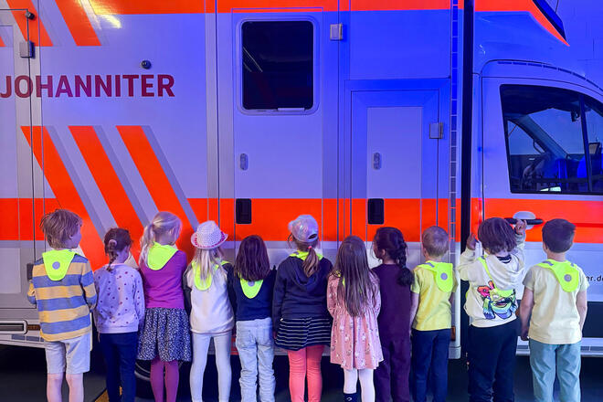 Kinder vor einem Rettungswagen. Die Fahrzeughalle ist in blaues Licht getaucht. 