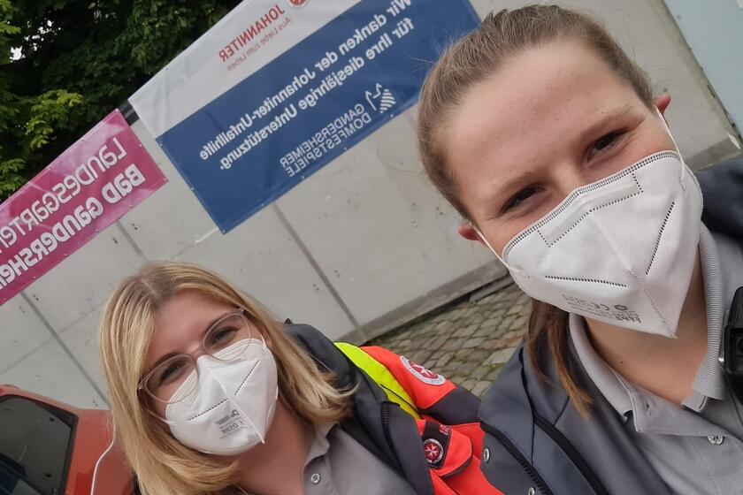 2 junge Frauen in Einsatzkleidung vor einem Dankesplakat an die Johanniter