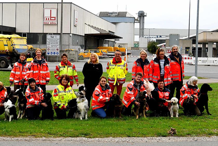 Gruppenfoto von Rettungshundeteams verschiedener Hilfsorganisationen mit ihren Hunden auf dem Gelände der Firma Max Bögl. Hundeführende in orangen und gelben Einsatzjacken knien gemeinsam mit ihren Hunden auf einer Wiese.