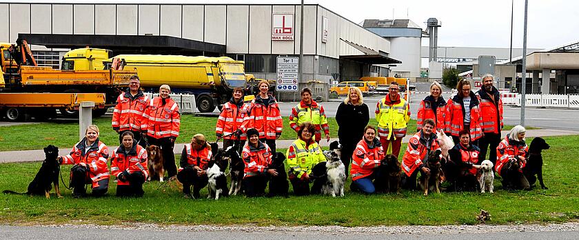Gruppenfoto von Rettungshundeteams verschiedener Hilfsorganisationen mit ihren Hunden auf dem Gelände der Firma Max Bögl. Hundeführende in orangen und gelben Einsatzjacken knien gemeinsam mit ihren Hunden auf einer Wiese.