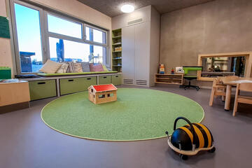 Gruppenraum der Kinderkrippe mit rundem Teppich, Sitzbank am Fenster und Spielzeug.