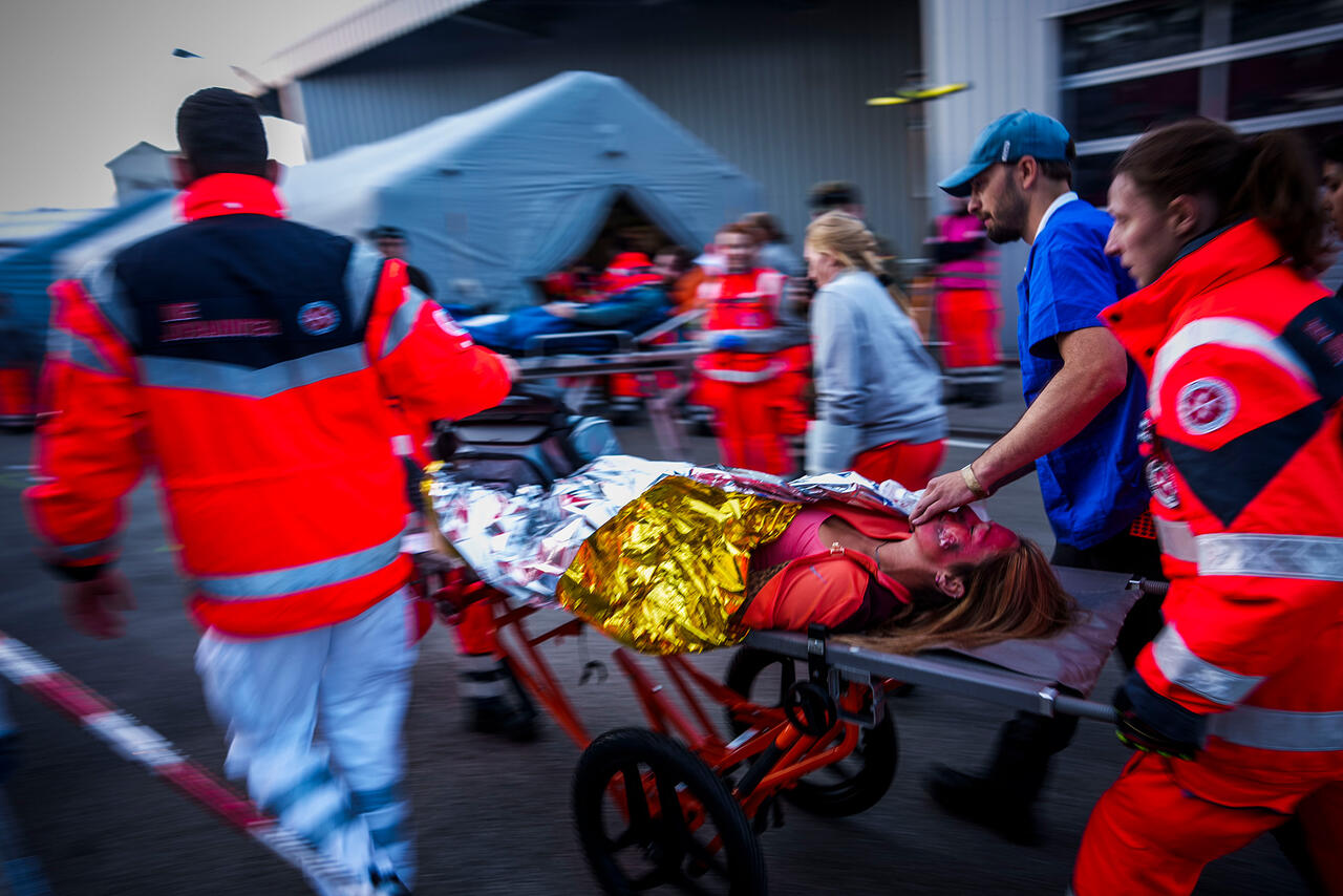 Rettungskräfte der Johanniter schieben eine Patientin auf einer Trage durch einen belebten Einsatzbereich; sie ist mit einer Rettungsdecke bedeckt, eine Person stabilisiert den Tubus, im Hintergrund steht ein Behandlungszelt.