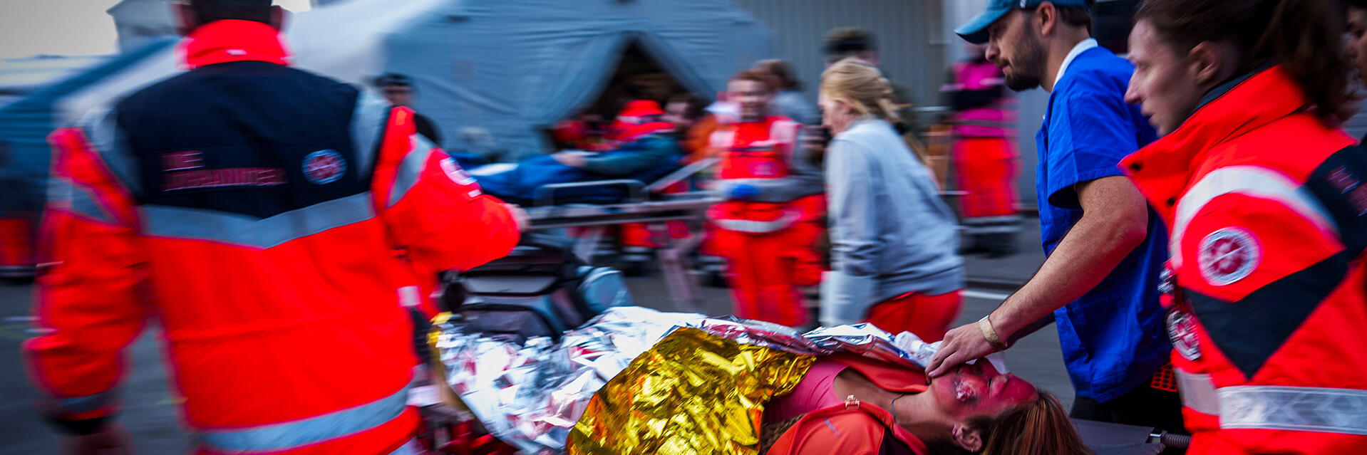 Rettungskräfte der Johanniter schieben eine Patientin auf einer Trage durch einen belebten Einsatzbereich; sie ist mit einer Rettungsdecke bedeckt, eine Person stabilisiert den Tubus, im Hintergrund steht ein Behandlungszelt.