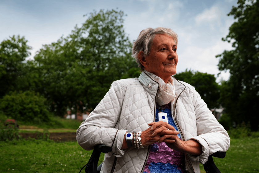 Ältere Frau mit kurzen grauen Haaren sitzt draußen auf einer Parkbank. Sie trägt eine helle Steppjacke und hält einen Johanniter-Hausnotruf-Sender sowie ein Hausnotruf-Armband. Im Hintergrund grüner Park mit Bäumen an einem bewölkten Tag. Die Frau blickt lächelnd und zufrieden in die Ferne.