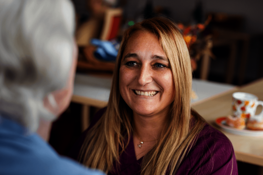 Eine Frau mit lange rötliche-blonde Haare lächelt eine Person mit grauen Haaren an, deren Hnterkopf angeschnitten und verschwommen von hinten zu sehen ist