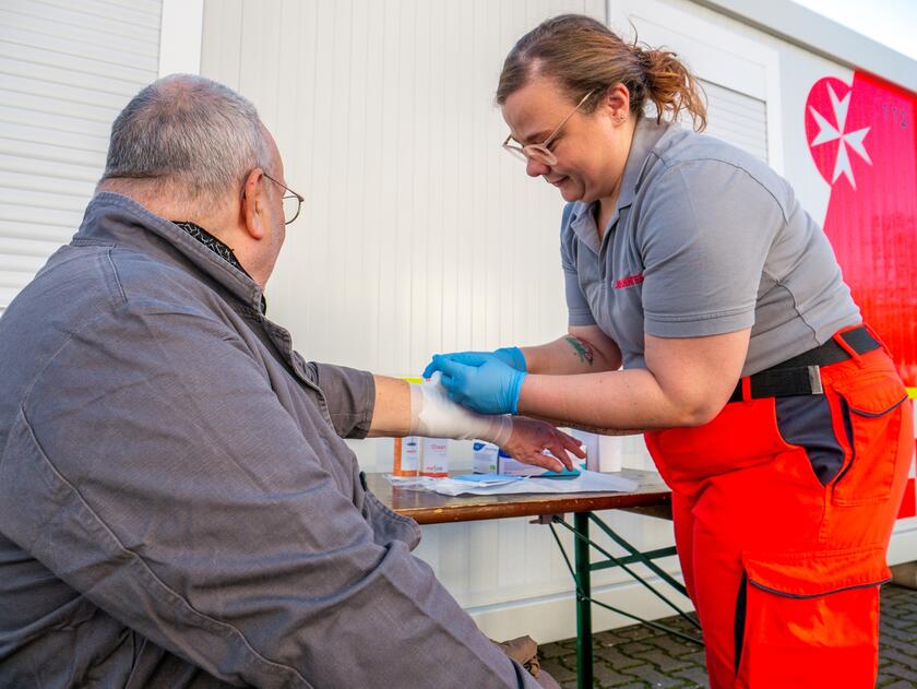 Eine Helferin der Johanniter beugt sich über den Arm eines älteren Mannes und legt einen Verband am Unterarm an.