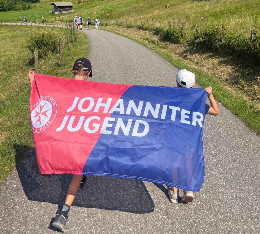 Jugendliche mit einer Johanniter-Jugend-Fahne auf dem Rücken schauen in Richtung Hütte