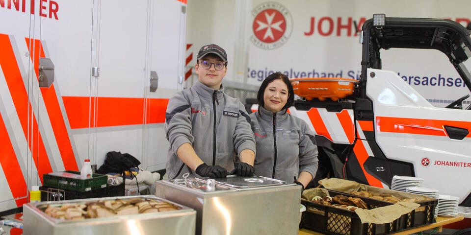 Ein junger Mann und eine junge Frau in Johanniter-Kleidung vor Tischen mit Essen darauf