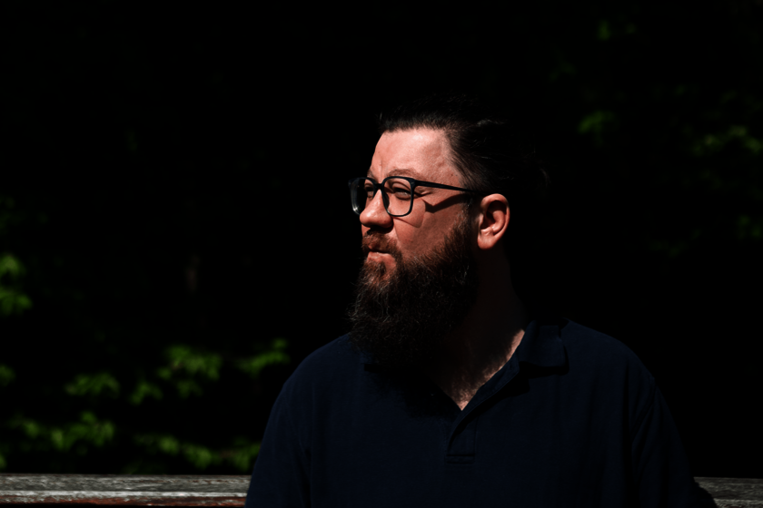 Ein bärtiger Mann im dunklen Poloshirt draußen, schaut nach rechts 