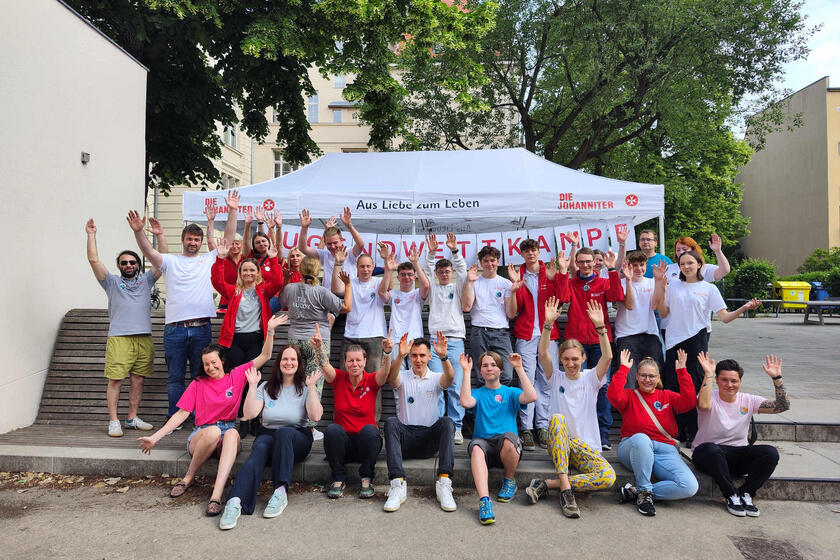 Eine größere Gruppe von Jugendlichen und Mitarbeitenden der Johanniter-Jugend posiert auf einer Treppe vor einem Pavillon für ein Foto und streckt die Hände nach oben.