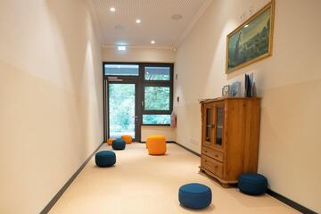 Ein gemütlicher Raum mit orangenen und blauen Sitzpolstern vor einem großen hellen Fenster.