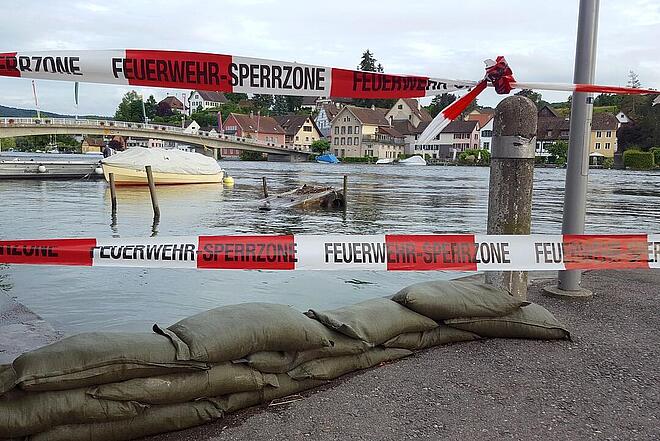 Ufer mit Sandsäcken und Absperrband „FEUERWEHR‑SPERRZONE“, teilversunkenes Wrack im Wasser, Brücke und Häuser im Hintergrund