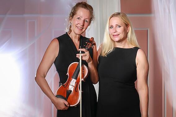 Die beiden Musikerinnen von Duo Virtuos stehen in schwarzer Konzertkleidung nebeneinander. Eine Dame hält ihre Violine in der Hand, im Hintergrund fällt warmes Licht durch transparente Vorhänge.