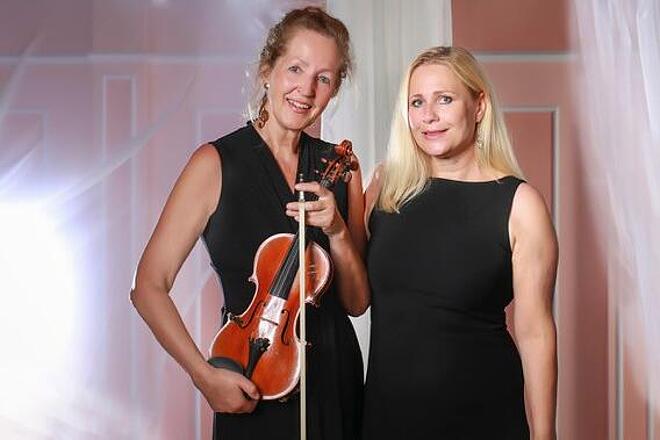 Die beiden Musikerinnen von Duo Virtuos stehen in schwarzer Konzertkleidung nebeneinander. Eine Dame hält ihre Violine in der Hand, im Hintergrund fällt warmes Licht durch transparente Vorhänge.