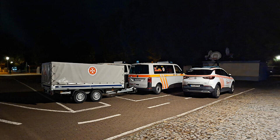 Johanniter-Fahrzeuge auf einem Parkplatz. Ein Mannschaftstransportwagen hat einen Anhänger. Es ist tiefste Nacht.