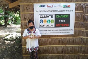 Ein Frau mit Mundschutz steht mit Pilzen in den Händen vor dem Projektbanner