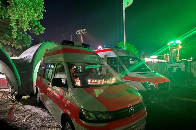 Zwei Rettungswagen stehen nachts nebeneinander auf einem Festivalgelände. Die Fahrzeuge sind beleuchtet, im Hintergrund ist ein großes Zelt zu sehen. Grüne Lichter tauchen die Szene in stimmungsvolles Licht, eine „First Aid“-Fahne weht über dem Einsatzbereich. Die Atmosphäre vermittelt Einsatzbereitschaft und Sicherheit während des Parookaville Festivals.