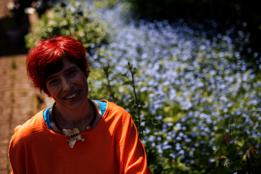 Eine Frau mit rotem Haar sitzt draußen vor blühendem Pflanzenbeet und lächelt