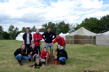 Eine Gruppe von jungen Menschen posiert mit Schaufel vor einem Loch auf einer Wiese. Im Hintergrund stehen Zelte
