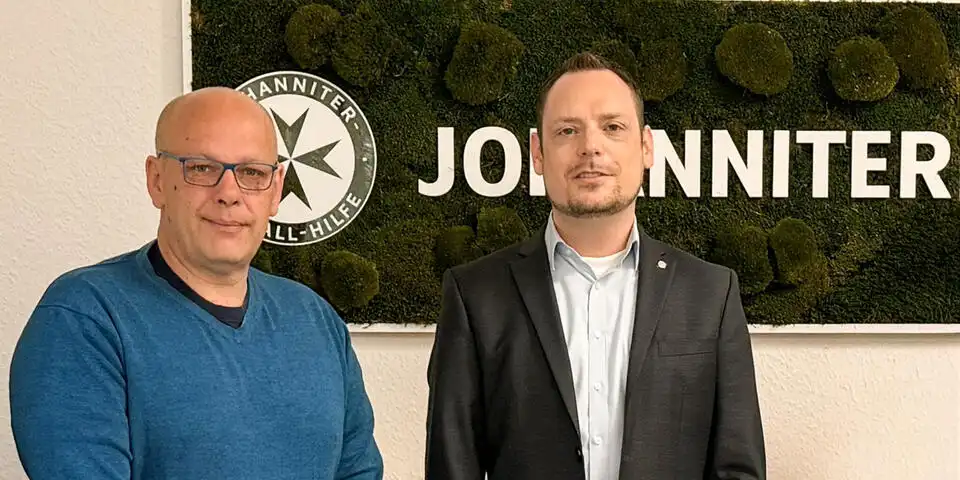 Zwei Männer schauen in die Kamera. Sie stehen vor einem Moosbild mit Johanniter-Logo