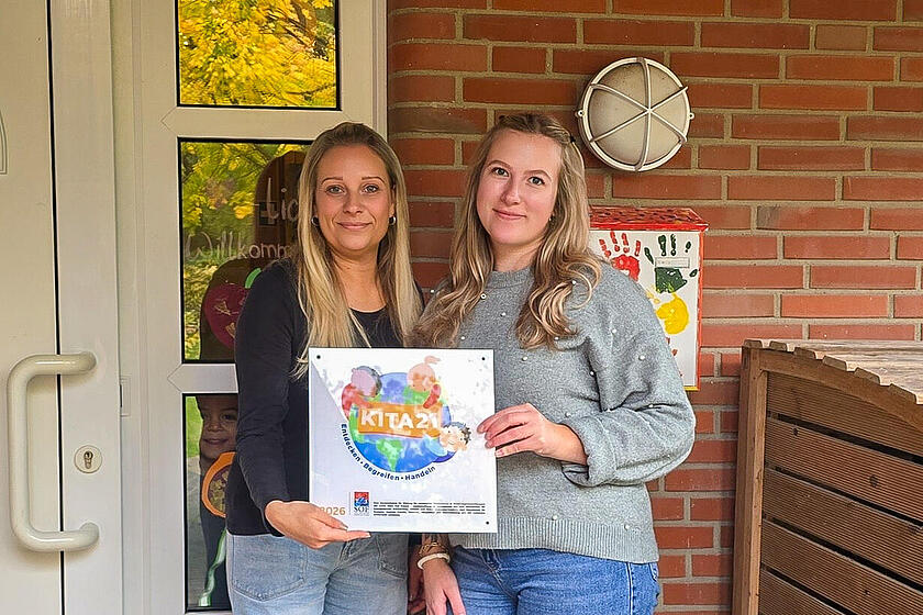 Andrea Kühn und Lilly Manke präsentieren stolz die KITA21-Plakette vor der Johanniter-Kita Pfützenracker.