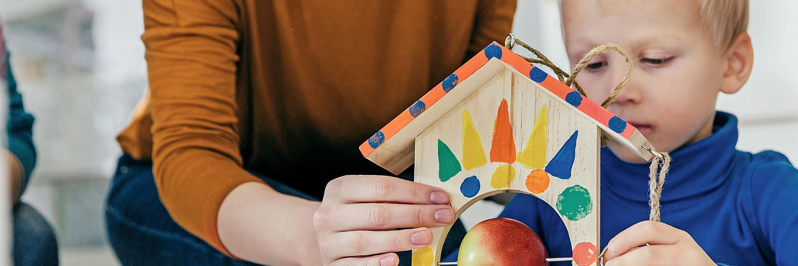 Eine erwachsene Person hilft einem kleinen Kind, ein bunt bemaltes Holz-Vogelhaus zu basteln. Das Kind hält das Häuschen fest, während darin ein Apfel auf einem Stab steckt. Beide wirken konzentriert und arbeiten gemeinsam am Tisch.
