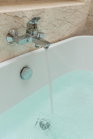 Ein silberner Wasserhahn ist voll aufgedreht. Das Wasser fließt in eine weiße Badewanne.