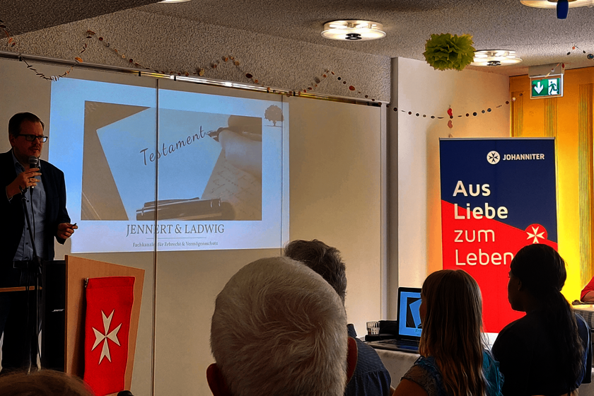 Links ein Mann im Anzug, er hält ein Mikrofon. Rechts einige Zuhörende von hinten zu sehen.