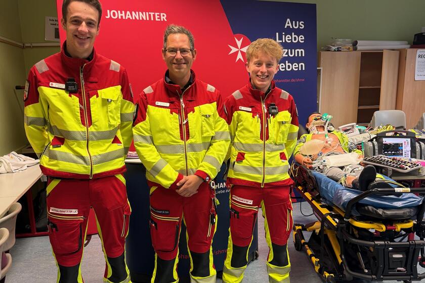 Drei Einsatzkräfte aus dem Rettungsdienst mit einer Rettungstrage und Puppe darauf