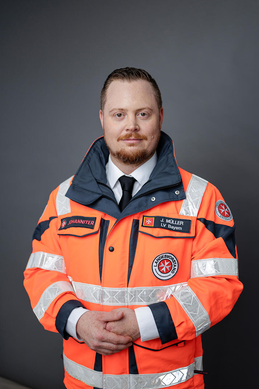 Porträt von Julian Müller in Johanniter-Einsatzjacke vor einem neutralen Hintergrund.