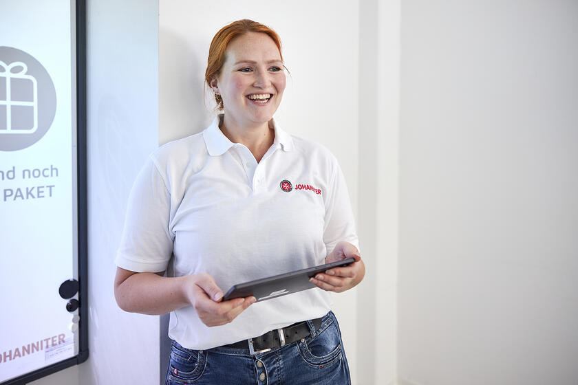 Johanniter-Mitarbeiterin im weißen Polohemd steht lächelnd mit Tablet in der Hand.