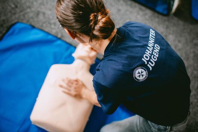 Man sieht eine junge Person von hinten oben. Sie kniet im Johanniter-Jugend-T-Shirt vor einer Erste-Hilfe-Puppe und drückt auf den Brustkorb.