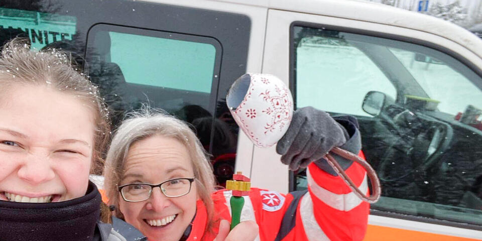 " Johanniterinnen lachen. Eine hält eine Kuhglocke hoch. Dahinter ein Johanniter-Fahrzeug in verschneiter Landschaft