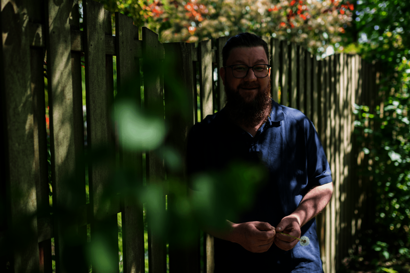 Ein Mann mit Bart lehnt an einem Holzzaun und hält eine Pusteblume in den Händen