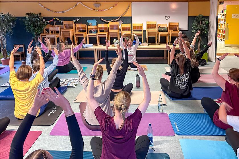 Eine Gruppe von Menschen macht Pilates: Alle sitzen und heben die Arme in die Luft.