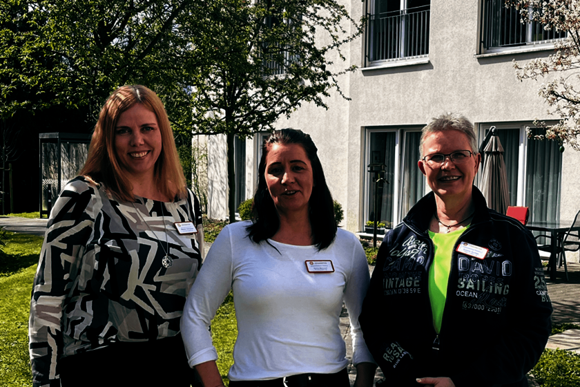 Drei Frauen stehen nebeneinander in einem frühlingshaften Garten vor einem weißgestrichenen Gebäude.