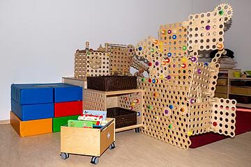 Spielbereich mit großem Holzgestell, das mit Spielzeugschrauben zusammengeschraubt ist. Davor steht ein kleiner Rollwagen aus Holz mit Kinderbüchern drinnen.