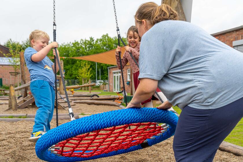 Eine Erzieherin stößt die Schaukel an, auf der zwei Kinder stehen.