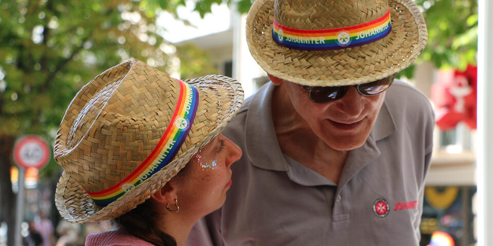 Zwei Menschen mit CSD-Strohhüten reden miteinander, er beugt sich runter um sie besser zu hören. im Hintergrund Bäume und Passantinnen und Passanten.