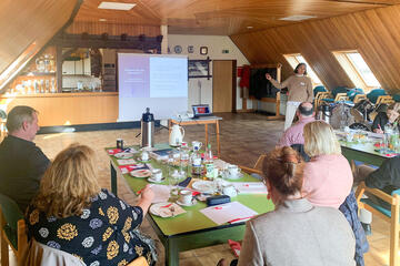 Im Gemeindehaus Tramm findet ein Grundlagenseminar statt. Zu sehen sind Ehrenamtliche, die an einem Tisch sitzen, und einer vortragenden Person vor einer Leinwand zuhören.