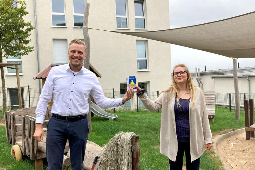 Dennis Schreiber (Vertreter der VGH in Northeim) übergibt die Kinderfinder an Tanja Schmidt (Einrichtungsleiterin der Kita am Wieter). Im Hintergrund sieht man die Außenspielgeräte der Kita. 