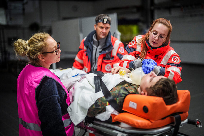 Johanniter-Rettungskräfte versorgen einen liegenden Patienten auf einer Trage in einem Innenraum; eine Helferin beatmet ihn mit einem Beutel, zwei Teammitglieder unterstützen.